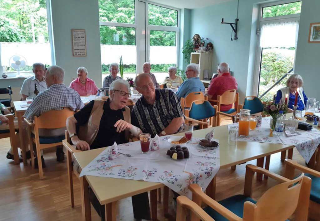 Eine Gruppe &auml;lterer Menschen sitzt in einem gem&uuml;tlichen Raum an Tischgruppen, offenbar bei einer Feier oder einem Gemeinschaftsessen.