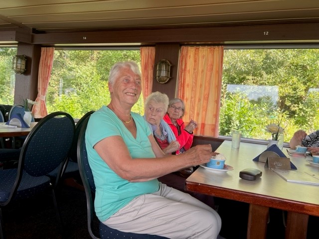 Drei ältere Personen sitzen entspannt an einem Tisch in einem Raum mit großen Fenstern.