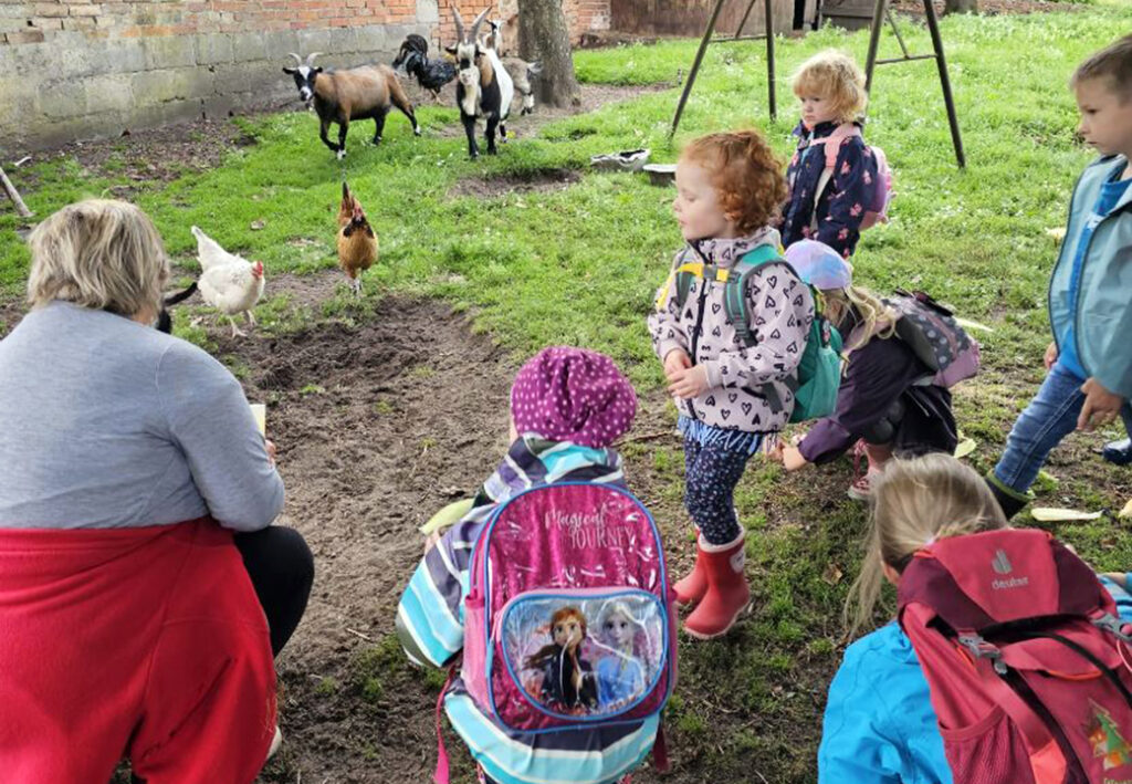 Mehrere Kinder mit Rucks&auml;cken stehen im Freien und beobachten H&uuml;hner und Ziegen.