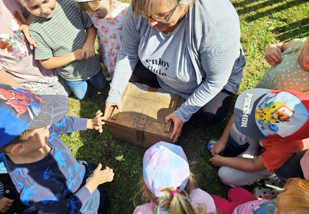 Eine Gruppe von Kindern um eine erwachsene Person, die eine Holzkiste h&auml;lt.