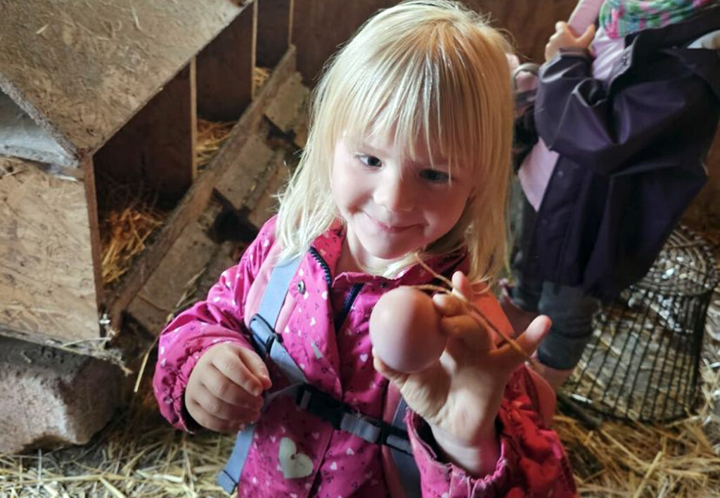 Ein Kind in einer pinkfarbenen Jacke steht in einem H&uuml;hnerstall und h&auml;lt ein Ei in der Hand.