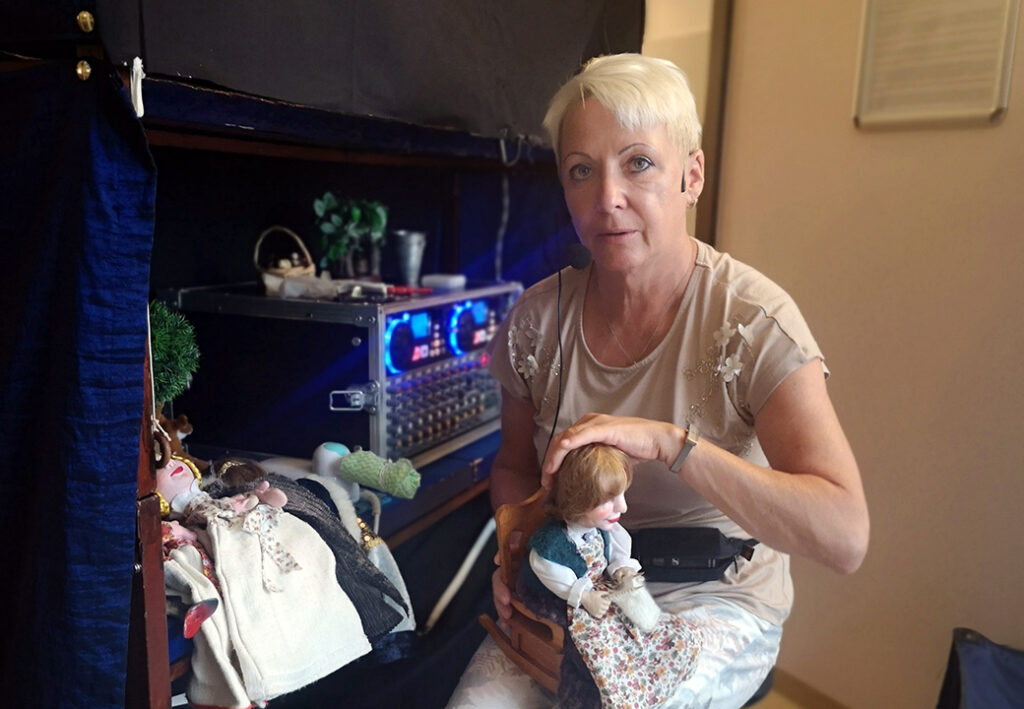 Eine Frau mit kurzem, blondem Haar sitzt neben einer B&uuml;hne mit Puppen.
