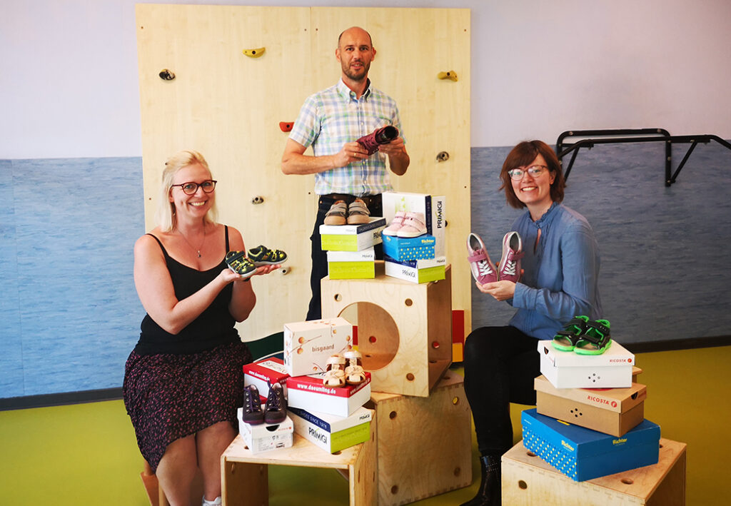 Drei Personen präsentieren Kinderschuhe vor einer Kletterwand.