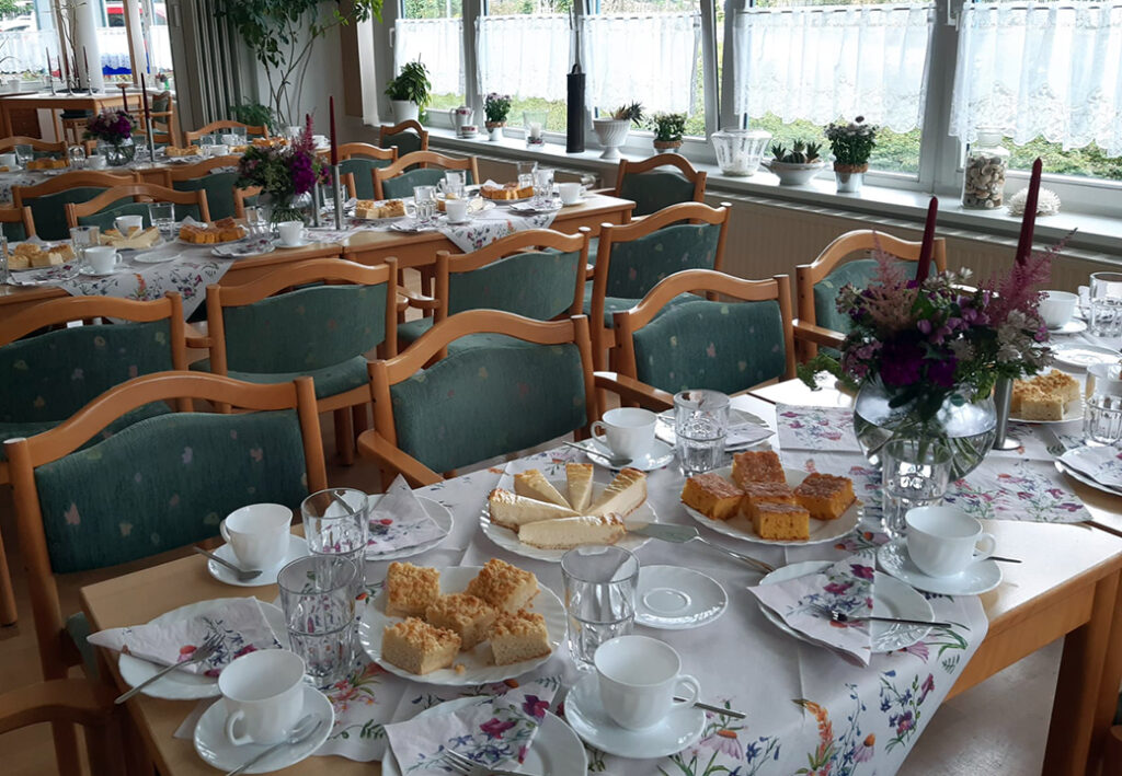 Gem&uuml;tlich gedeckter Tisch mit Kuchen und Kaffeegeschirr in einem Raum mit mehreren Tischen und St&uuml;hlen.