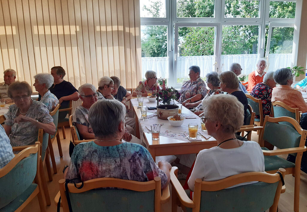 Gruppe &auml;lterer Menschen bei einer Versammlung in einem Raum mit gro&szlig;en Fenstern.