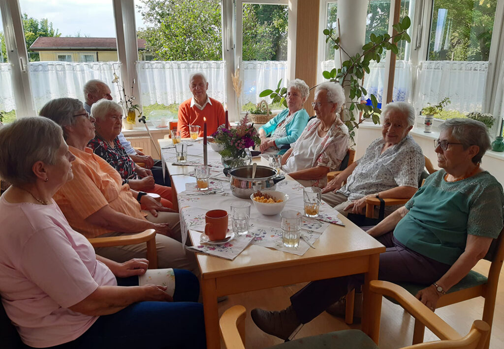 Gruppe &auml;lterer Menschen sitzt in einem gem&uuml;tlichen Raum um einen gedeckten Tisch.