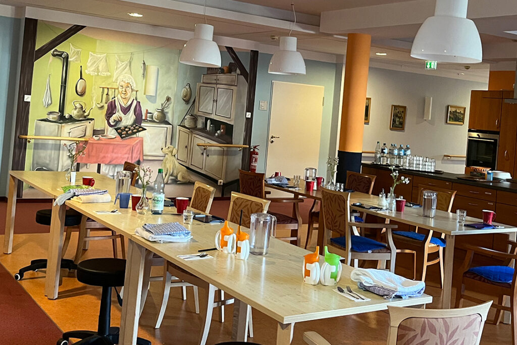 Ein Gemeinschaftsraum mit Tischen und Stühlen, Wandbild einer Küche im Hintergrund.