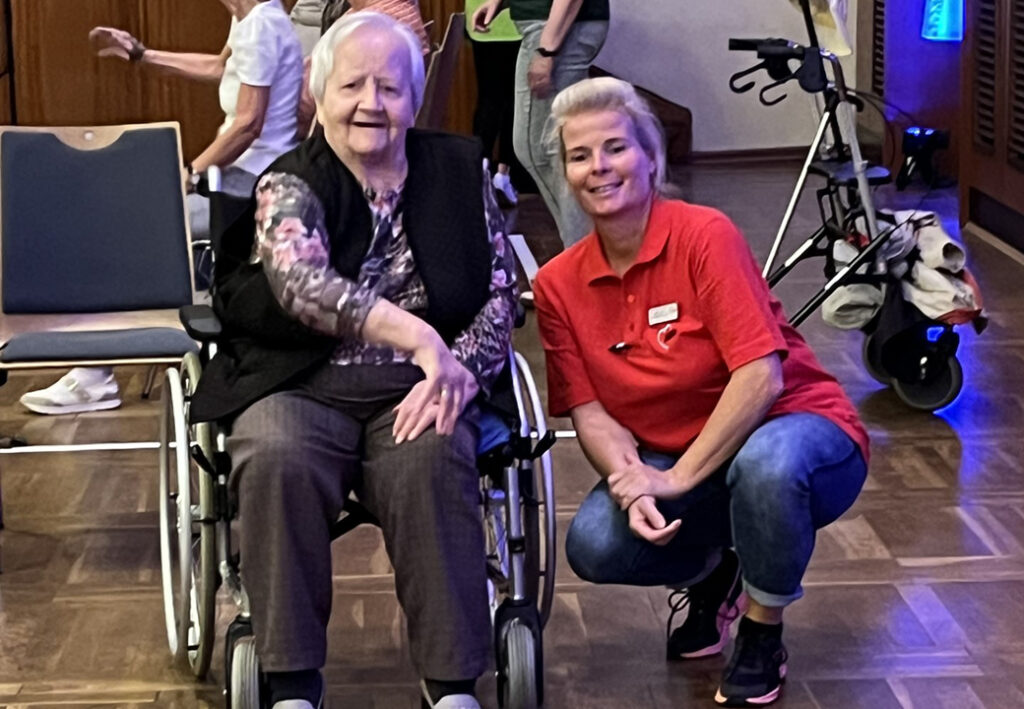 Eine &auml;ltere Frau sitzt in einem Rollstuhl, neben ihr kniet eine j&uuml;ngere Frau in rotem Shirt.