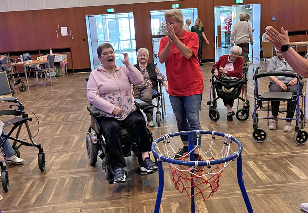 Mehrere &auml;ltere Personen bei einem Spiel mit einem mobilen Basketballkorb in einem Gemeinschaftsraum.