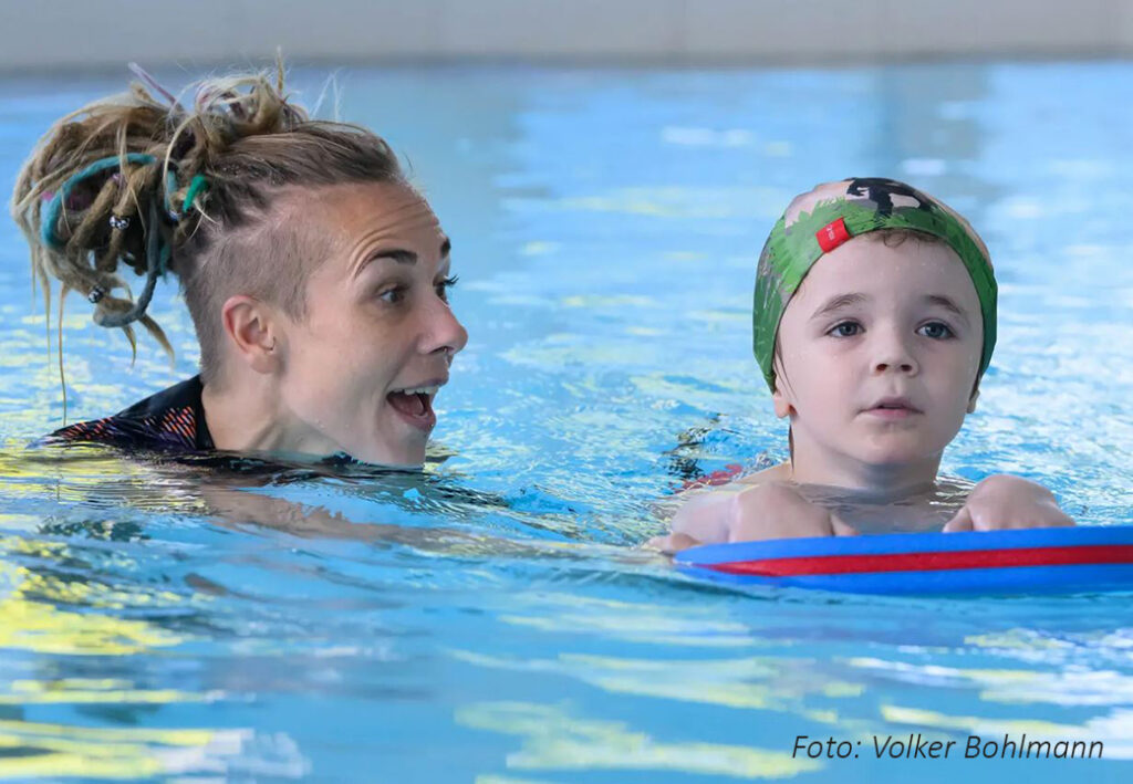 Erwachsene mit bunter Frisur im Schwimmbecken neben Kind mit Schwimmbrett