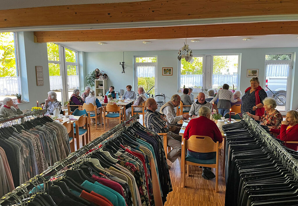 Raum mit mehreren &auml;lteren Menschen, die an Tischen in einem Gemeinschaftsraum sitzen und essen, mit Kleiderst&auml;ndern im Vordergrund.