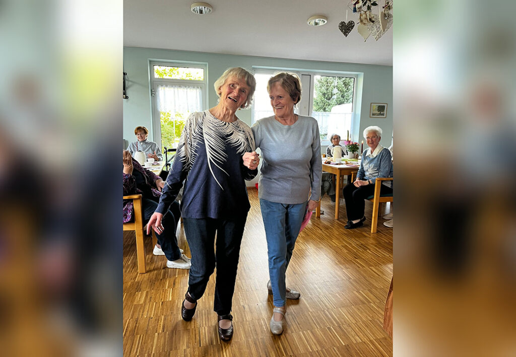 Zwei &auml;ltere Frauen tanzen gemeinsam in einem Raum mit anderen Senioren.