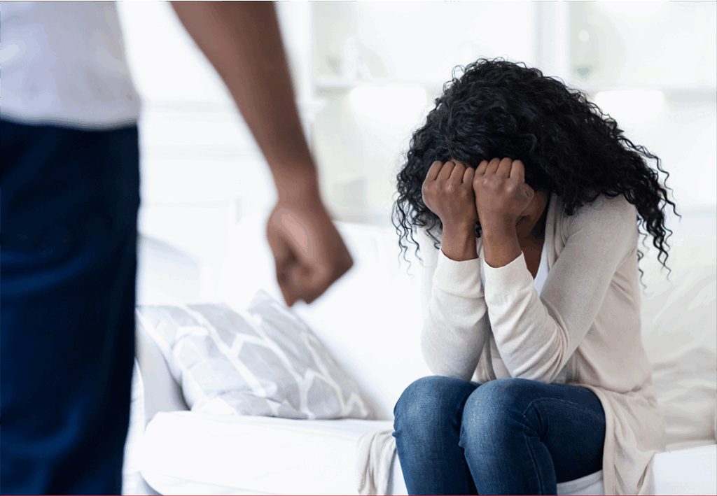 Eine Frau sitzt mit gesenktem Kopf auf einem Sofa und hält die Hände vor das Gesicht, während eine stehende Person mit geballter Faust im Vordergrund sichtbar ist.