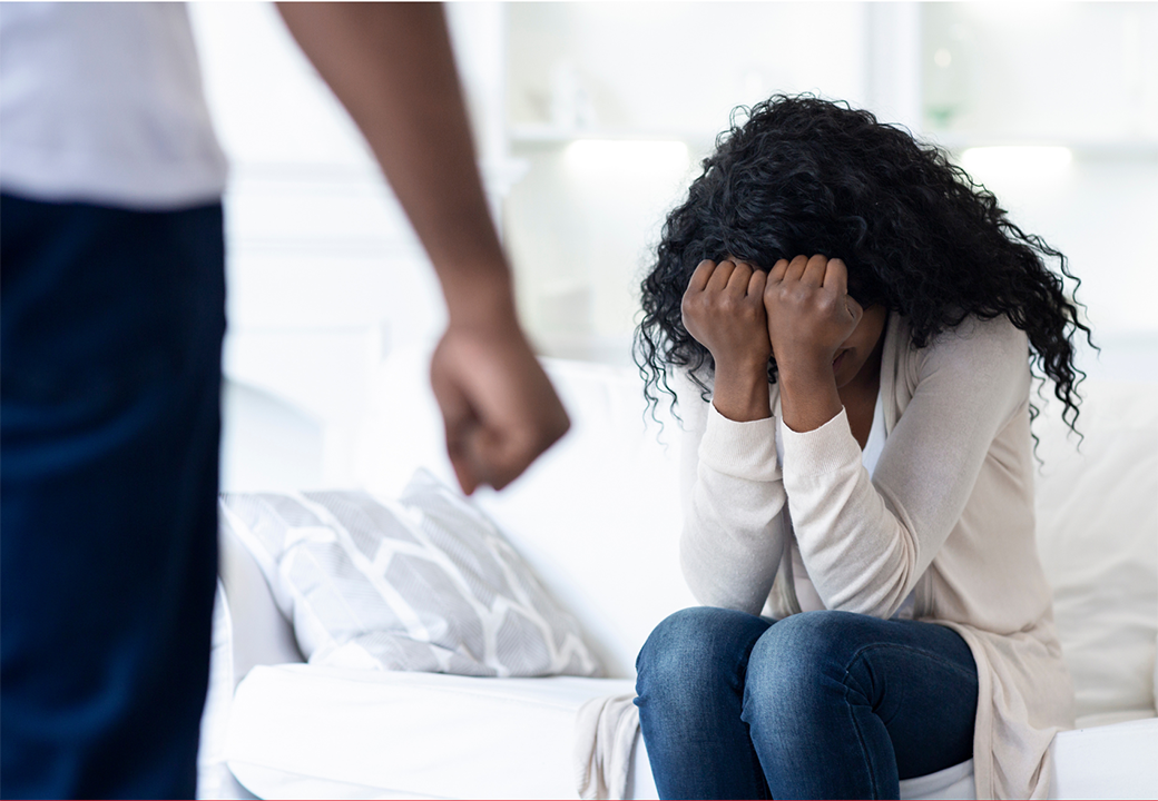 Eine Frau sitzt mit gesenktem Kopf auf einem Sofa und hält die Hände vor das Gesicht, während eine stehende Person mit geballter Faust im Vordergrund sichtbar ist.
