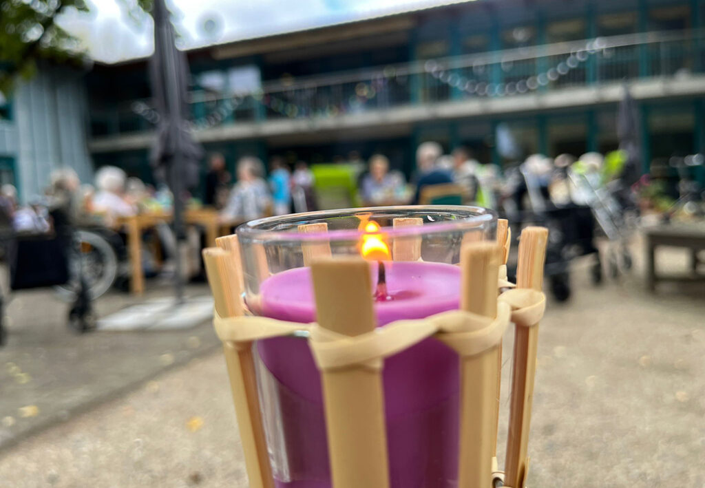 Ein lila Kerze brennt in einem Glasgef&auml;&szlig; im Vordergrund, w&auml;hrend im Hintergrund unscharf Menschen an Tischen vor einem Geb&auml;ude sitzen.