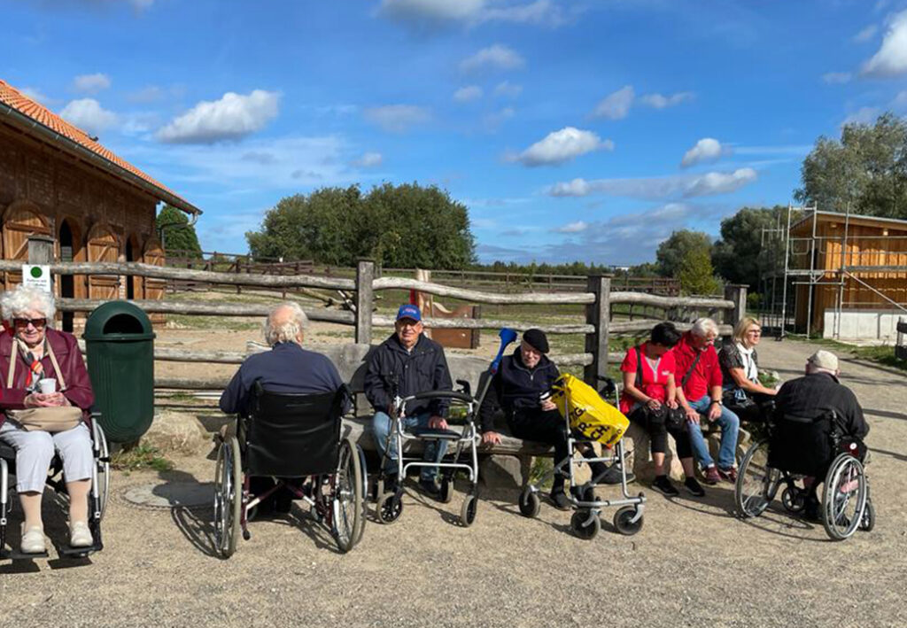 Eine Gruppe von &auml;lteren Menschen sitzt drau&szlig;en auf B&auml;nken und in Rollst&uuml;hlen, umgeben von einem h&ouml;lzernen Zaun und Geb&auml;uden im Hintergrund.