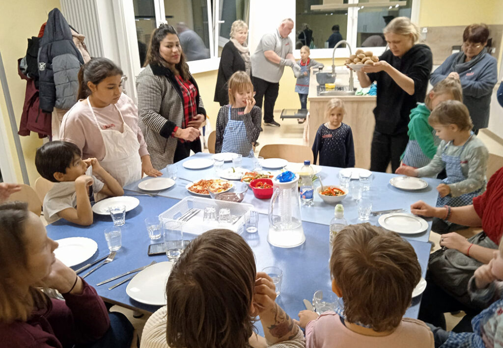 A group of people, including children, are gathered around a dining table set with plates and food.