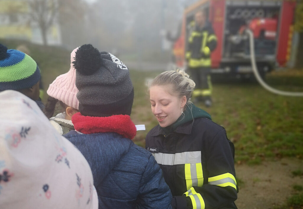 Eine Person in Einsatzkleidung der Feuerwehr spricht mit einer Gruppe von Kindern im Freien.