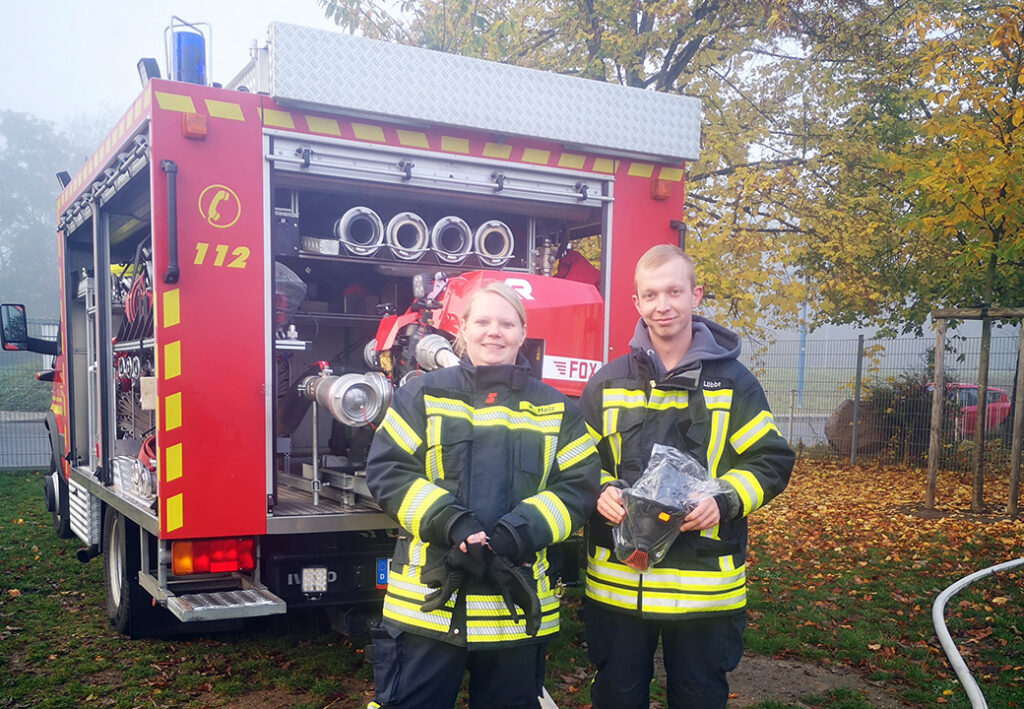 Zwei Feuerwehrleute in Einsatzkleidung vor einem Feuerwehrfahrzeug.