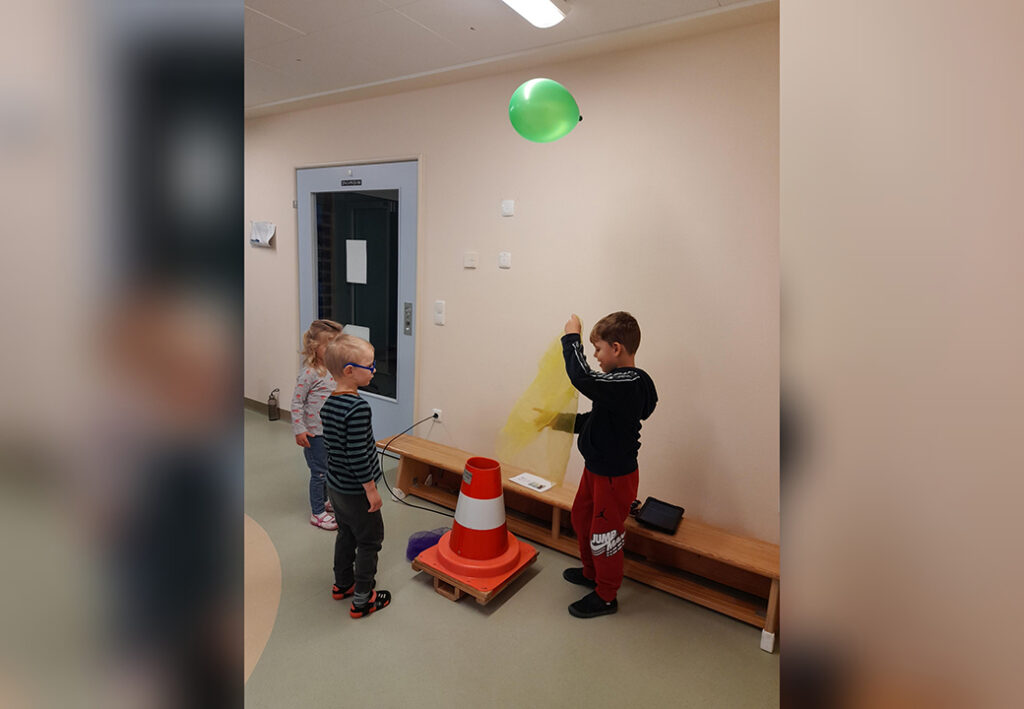 Drei Kinder in einem Flur, ein Junge h&auml;lt ein Blatt Papier unter einen schwebenden gr&uuml;nen Ballon