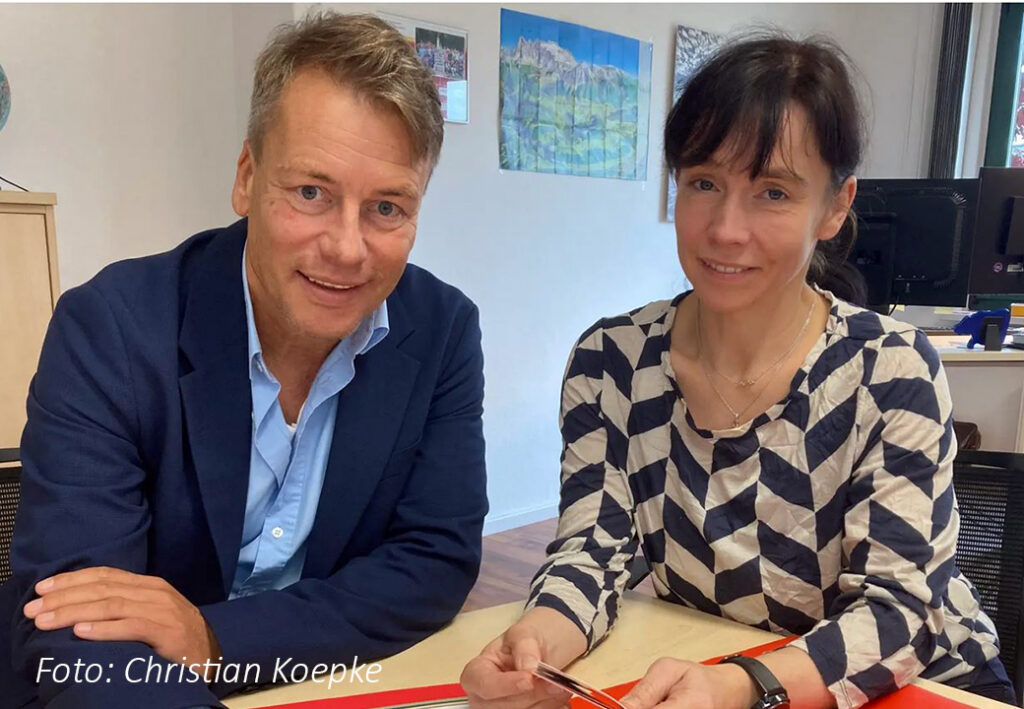 Foto von zwei Personen am Tisch in einem Büro, lächelnd und freundlich schauend.