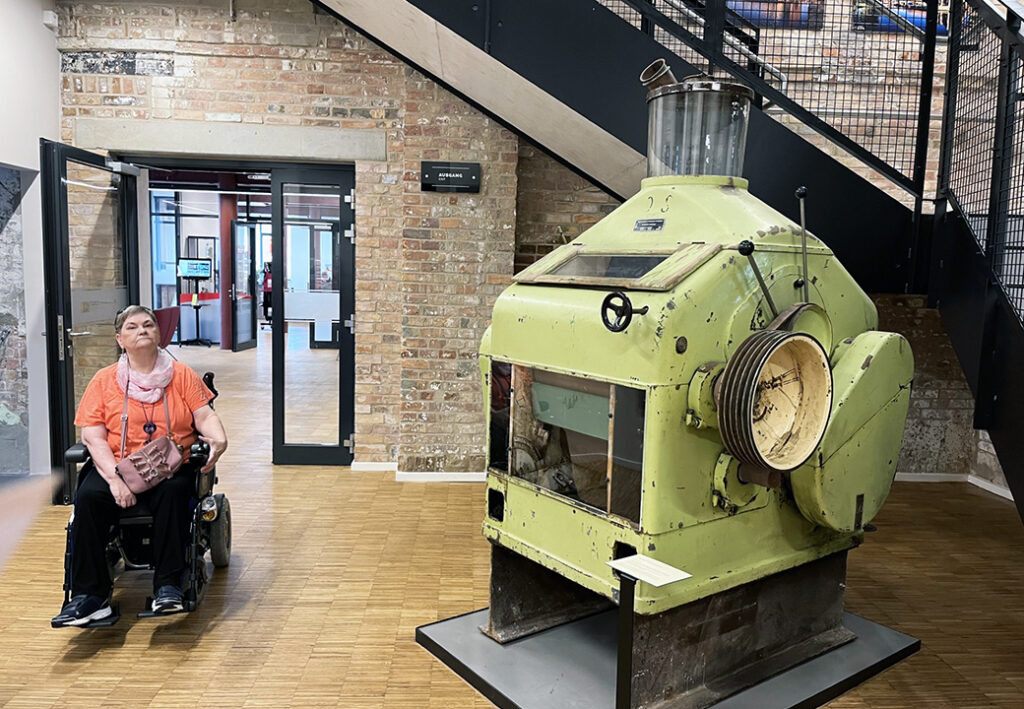 Eine Person in einem Rollstuhl vor einer alten, großen Maschine in einem Gebäude mit Backsteinwänden.