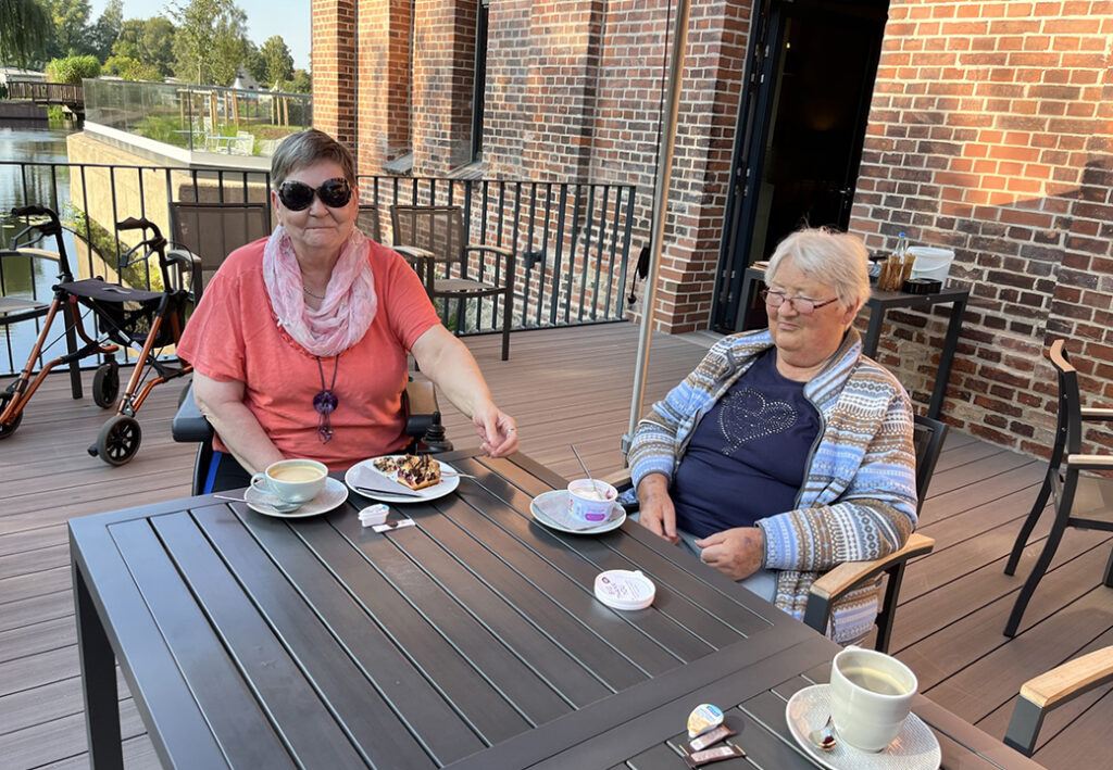 Zwei &auml;ltere Damen sitzen an einem Tisch auf einer Terrasse und genie&szlig;en Kaffee und Kuchen.
