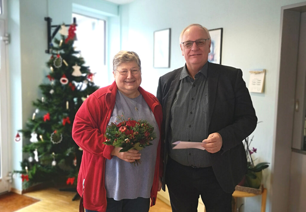 Zwei Personen posieren lächelnd, eine hält einen Blumenstrauß, die andere ein Blatt Papier. Im Hintergrund ein dekorierter Weihnachtsbaum.