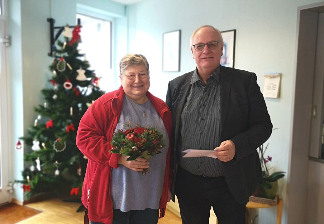 Zwei Personen posieren lächelnd, eine hält einen Blumenstrauß, die andere ein Blatt Papier. Im Hintergrund ein dekorierter Weihnachtsbaum.