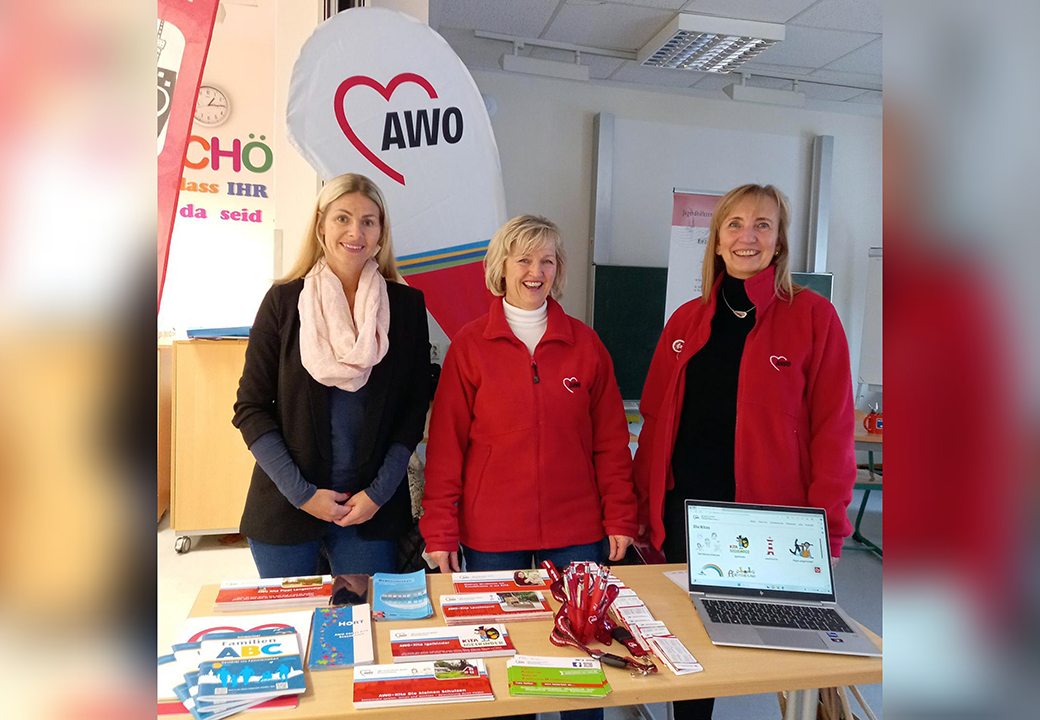Drei Frauen stehen hinter einem Tisch mit Broschüren und einem Laptop. Sie tragen AWO-Jacken und lächeln.