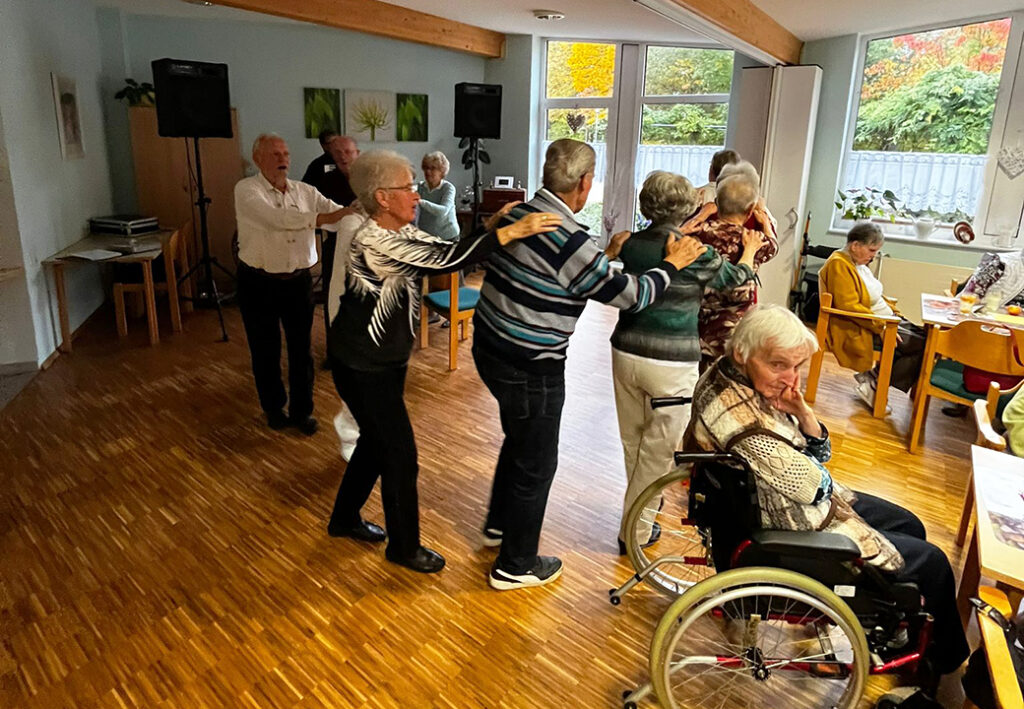 &Auml;ltere Menschen tanzen in einem Gemeinschaftsraum und bilden eine Polonaise.