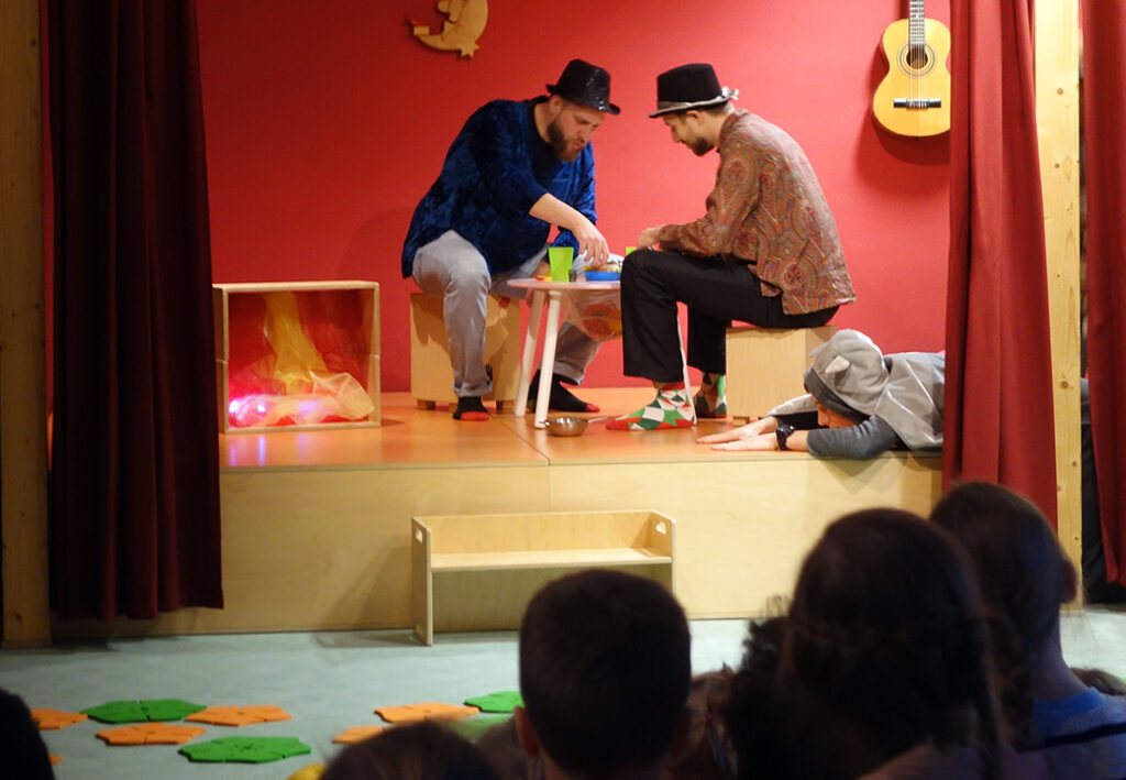 Drei Menschen in bunten Kost&uuml;men auf einer B&uuml;hne, eine Gitarre h&auml;ngt an der Wand.