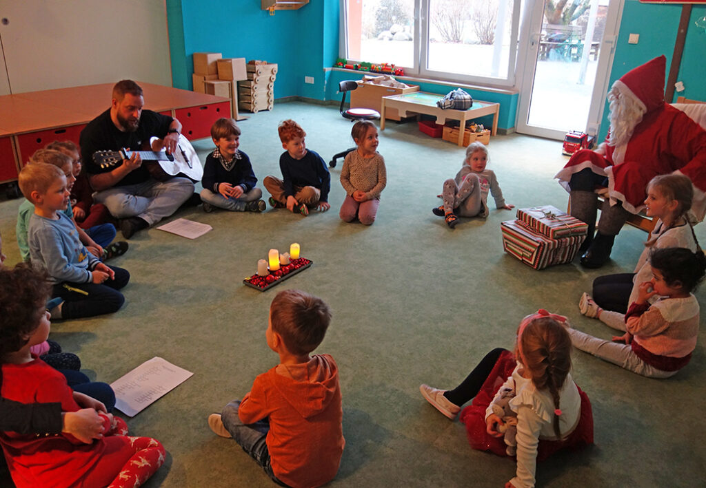 Kindergruppe sitzt im Kreis mit einem Mann, der Gitarre spielt, und einer als Weihnachtsmann verkleideten Person