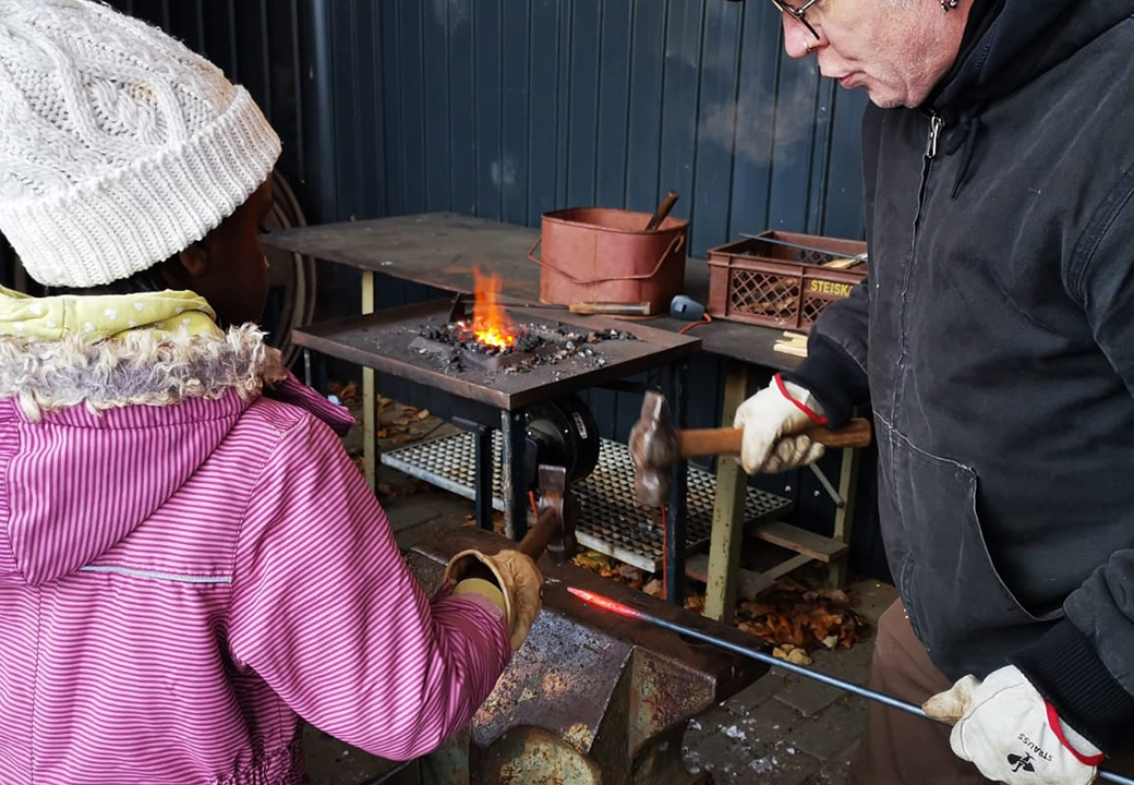 Ein Kind und ein Erwachsener schmieden gemeinsam ein Eisenstück an einem kleinen Amboss in einer Werkstatt mit Feuerstelle im Hintergrund.