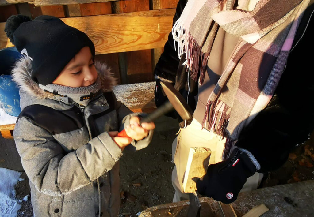 Ein Kind in Winterkleidung bearbeitet mit einem Erwachsenen zusammen ein Holzst&uuml;ck mit Hammer und Mei&szlig;el.