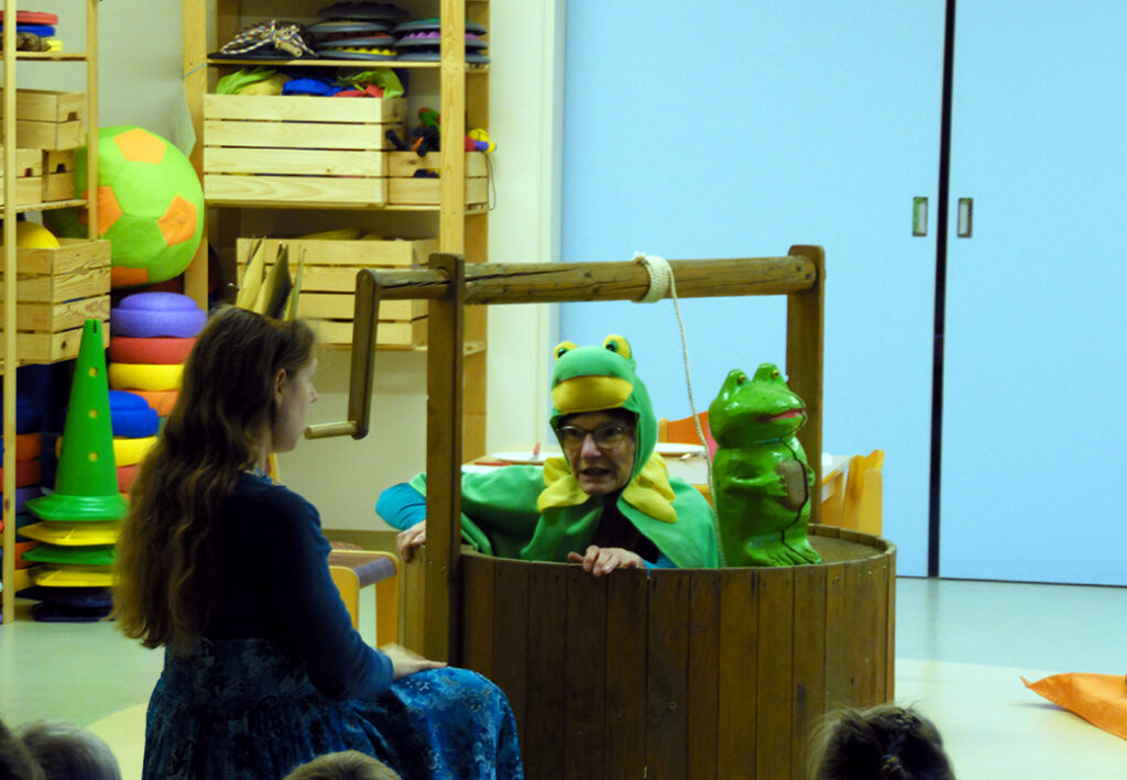 Kindertheaterauff&uuml;hrung in einem Raum mit Holzkisten voller bunter Spielsachen.