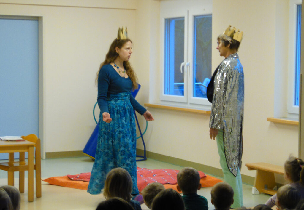 Zwei verkleidete Personen f&uuml;hren ein Theaterst&uuml;ck f&uuml;r Kinder auf, eine Person tr&auml;gt einen glitzernden Umhang und eine Krone, die andere ein langes blaues Kleid und ebenfalls eine Krone.