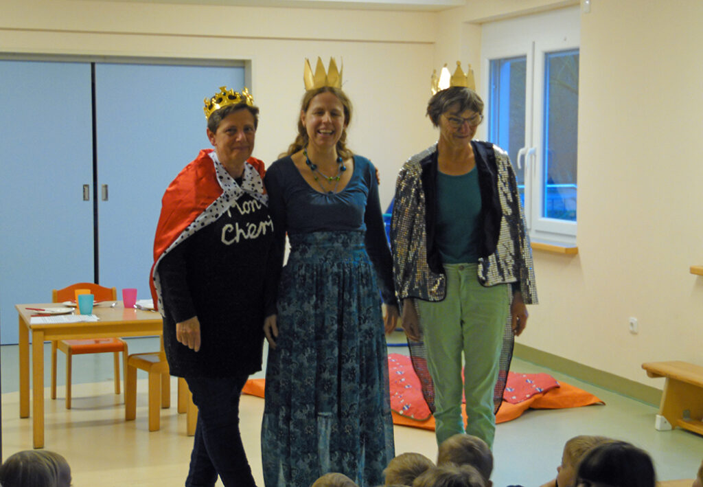 Drei Erwachsene tragen Kronen und Kleider und stehen in einem Raum mit Kindern.