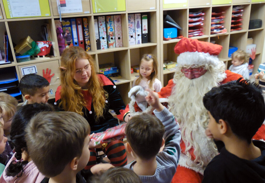 Kinder versammeln sich um einen als Weihnachtsmann verkleideten Erwachsenen und eine Frau in einem weihnachtlichen Kost&uuml;m, um ein Geschenk zu &uuml;berreichen.