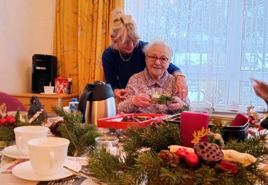 Eine &auml;ltere Frau, die von einer j&uuml;ngeren Frau umarmt wird, h&auml;lt ein Kerzenlicht in der Hand. Sie sitzen an einem Tisch mit Weihnachtsdekoration.