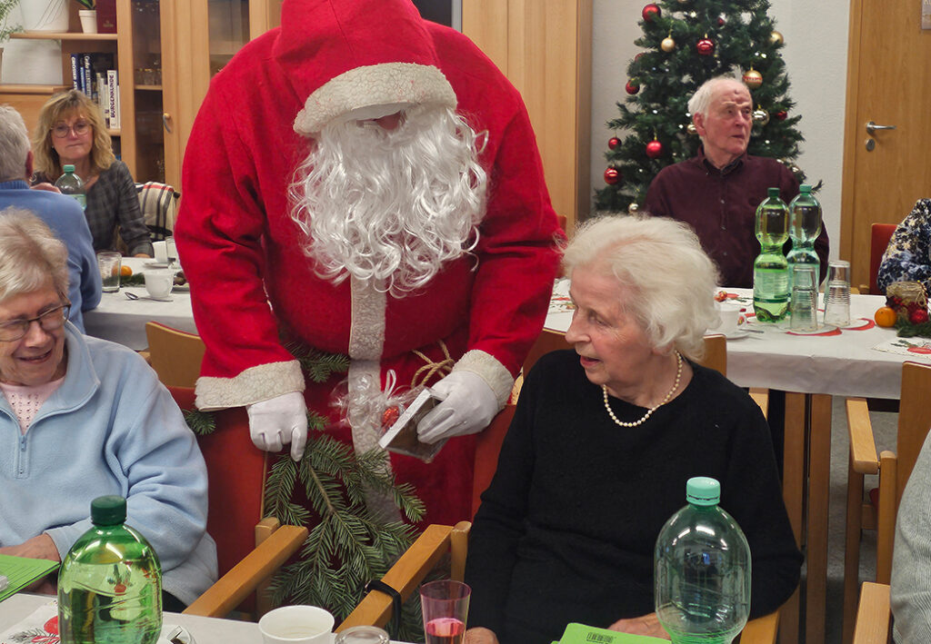 Eine Person im Weihnachtsmann-Kostüm verteilt Geschenke an eine ältere Frau in einem Gemeinschaftsraum.