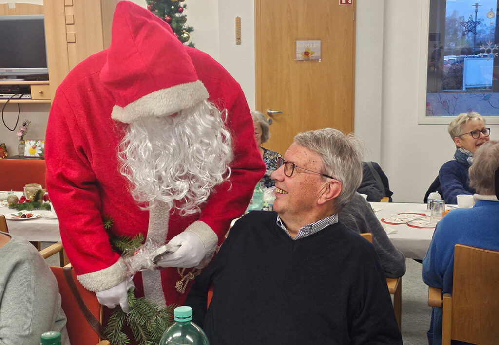 Ein Weihnachtsmann in rotem Kost&uuml;m spricht mit einem &auml;lteren Mann, der an einem Tisch sitzt.