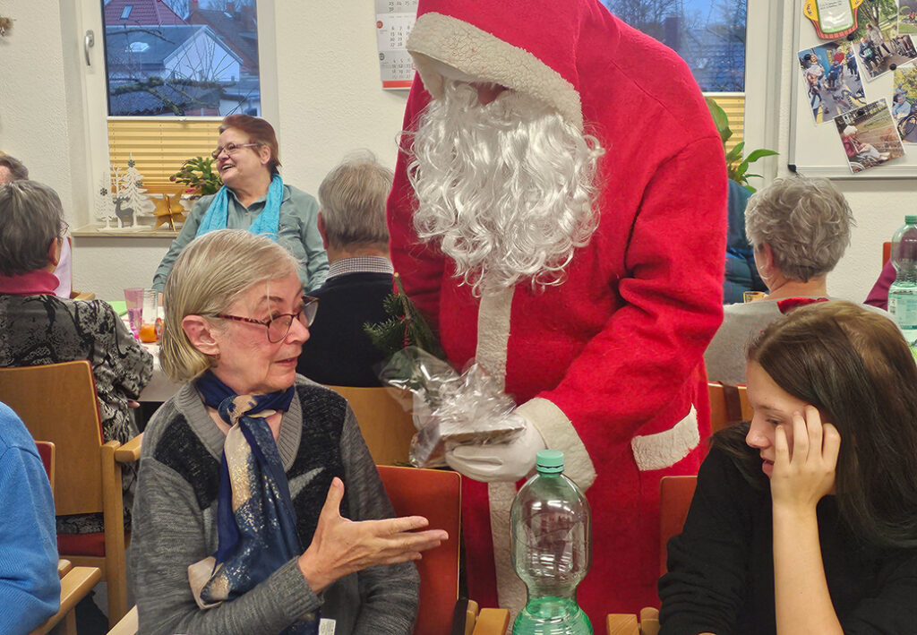 Weihnachtsmann schenkt einer &auml;lteren Frau ein Geschenk in einem Gemeinschaftsraum