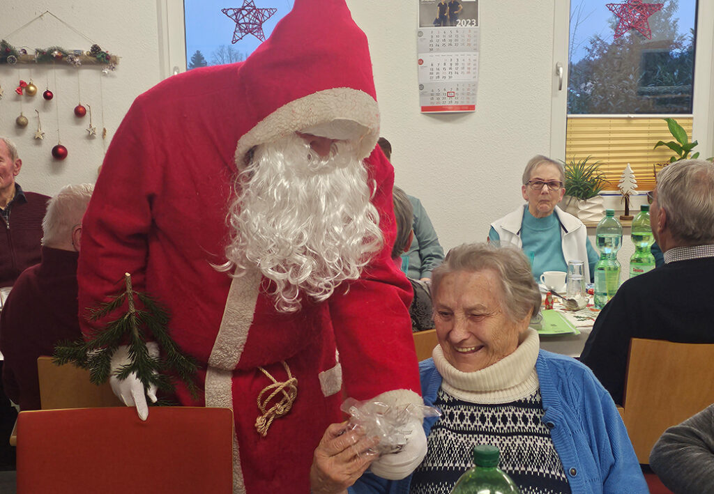 Person dressed as Santa Claus giving a gift to an elderly woman in a festive setting.