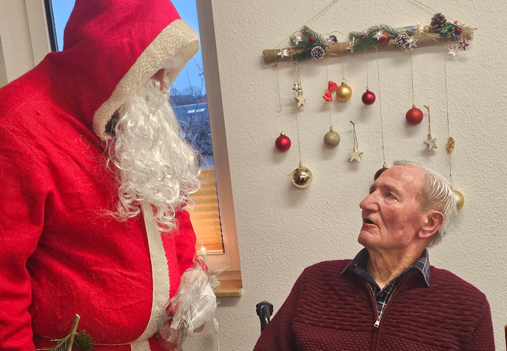 Ein als Weihnachtsmann verkleideter Mann steht neben einem &auml;lteren Mann, der sitzt.