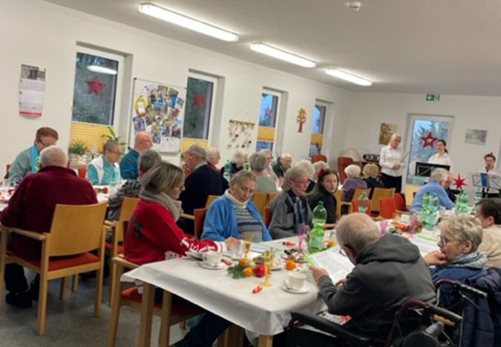 Eine Gruppe von Menschen sitzt in einem Gemeinschaftsraum an Tischen, die festlich gedeckt sind.