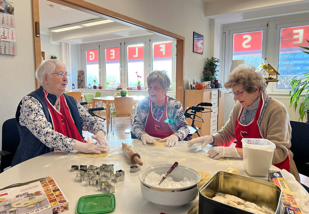 Drei ältere Frauen beim gemeinsamen Backen an einem Tisch in einem Gemeinschaftsraum.