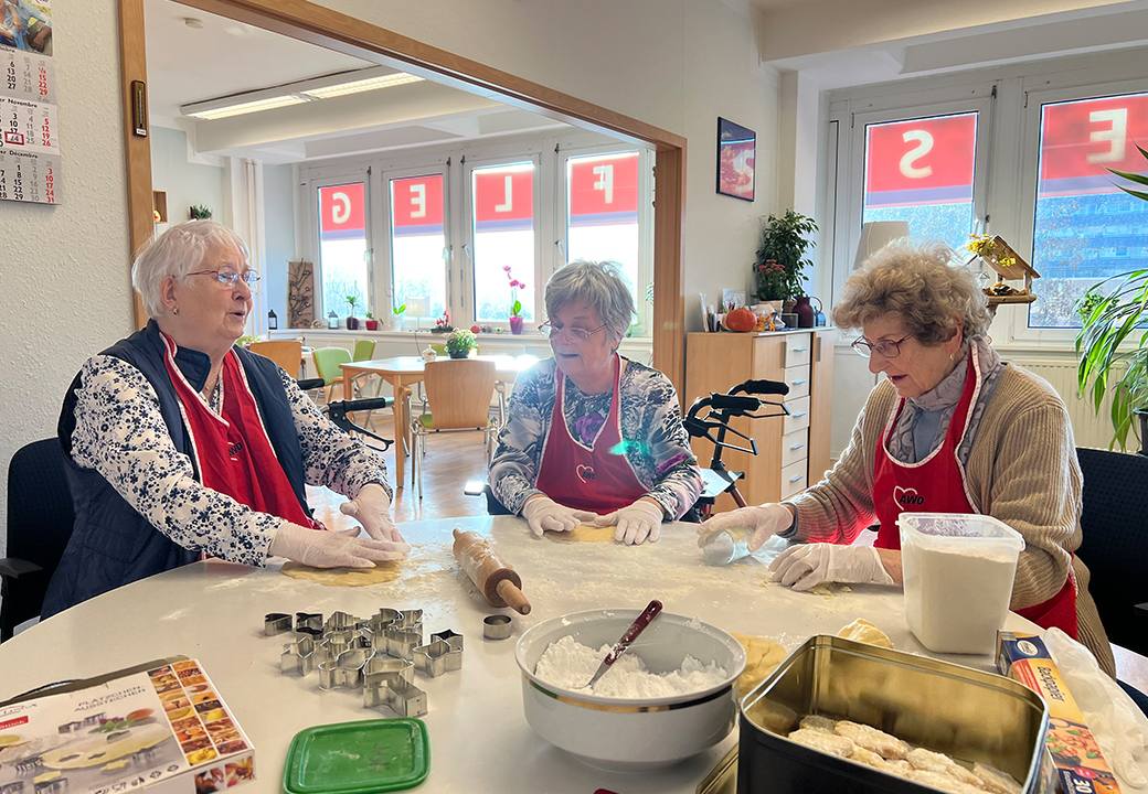 Drei ältere Frauen beim gemeinsamen Backen an einem Tisch in einem Gemeinschaftsraum.