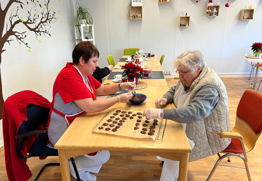 Zwei Personen, die gemeinsam Kekse an einem Tisch in einem hellen Raum zubereiten.
