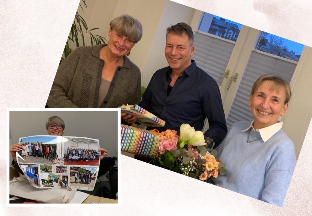 Drei Personen in einem Raum, eine ältere Frau, ein Mann und eine Frau, präsentieren Geschenke und Blumen.